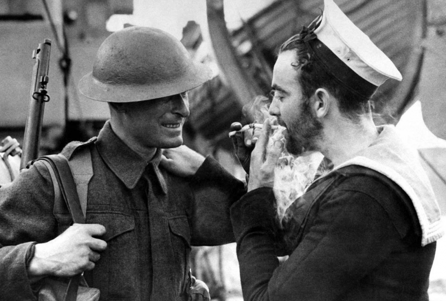 Два примера британских военных сил, солдат в боевой одежде и бородатый канадский моряк освещают огонь в английском порту 14 января 1941 года.